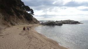 dos nois que caminen per la platja i arriben cap a unes roques planes que estan a sobre el mar
