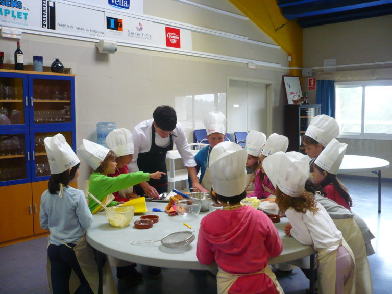 taula amb molts nens amb barrets de cuiner fent un taller de cuina