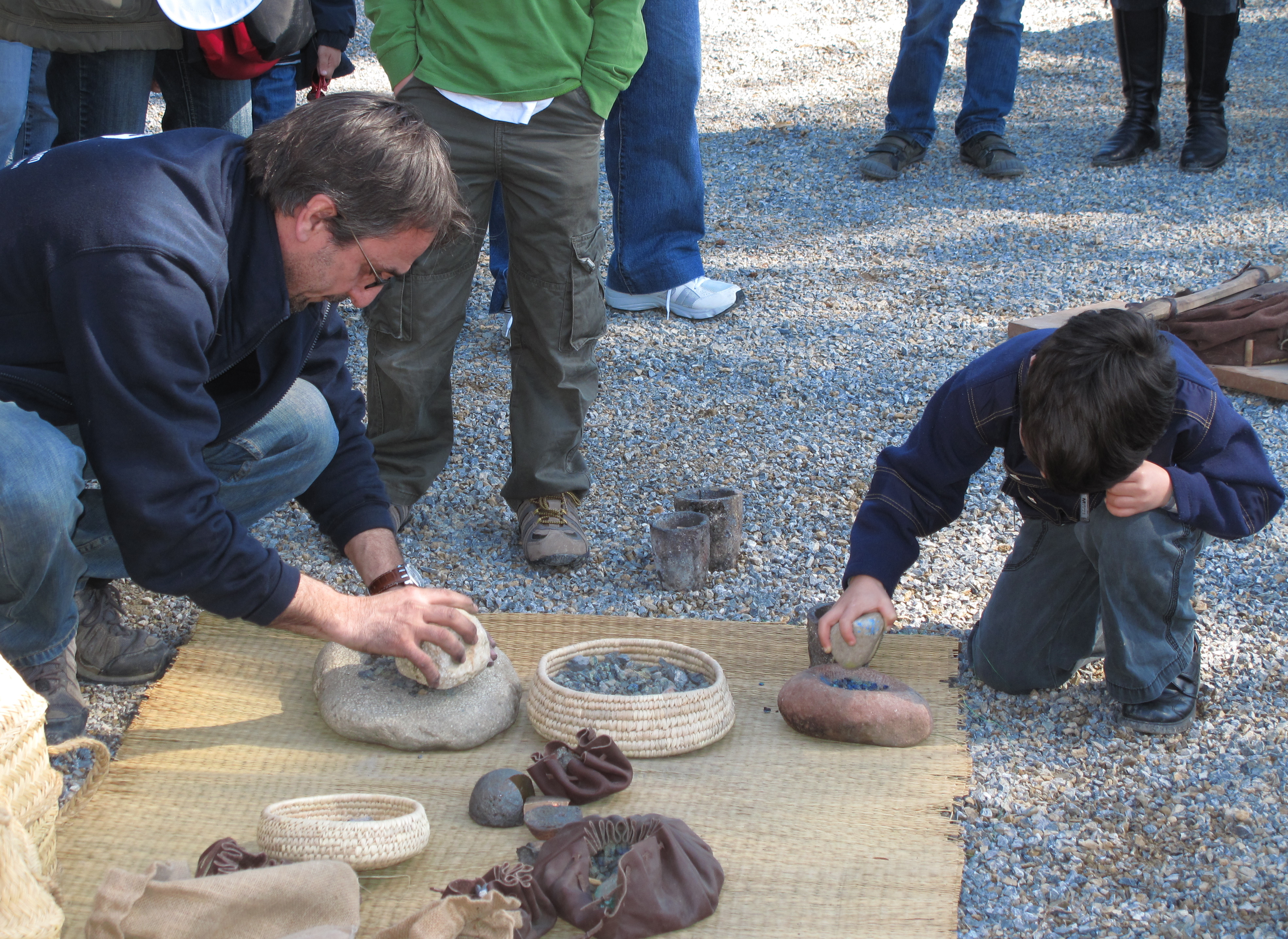 Taller amb pedres. Arqueòleg i un nen que pica unes pedres. Estan tots dos a terra i gent que els mira com ho fan