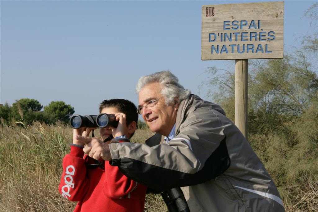 avi amb un nen. El nen mira amb uns binocles. A darrera seu hi ha un cartell de fusta que posa espai d'interès natural.