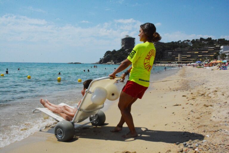 socorrista que acompanya a una nena amb una cadira que no és del tot amfíbia però que l'ajuda a estar a peu de sorra de la platja amb l'aigua just davant seu