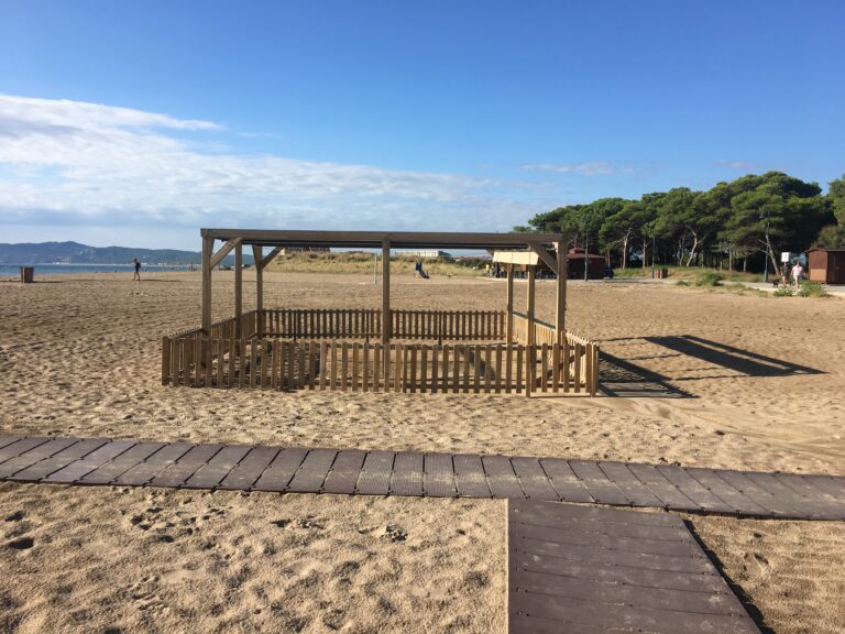 socorrista que acompanya a una nena amb una cadira que no és del tot amfíbia però que l'ajuda a estar a peu de sorra de la platja amb l'aigua just davant seu