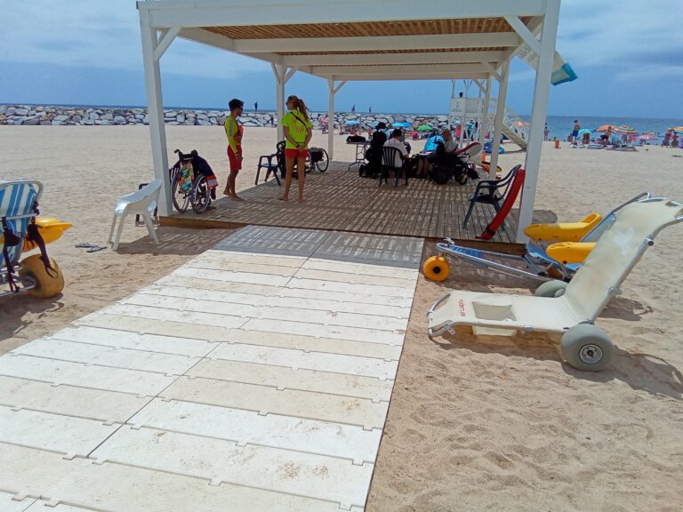 socorrista que acompanya a una nena amb una cadira que no és del tot amfíbia però que l'ajuda a estar a peu de sorra de la platja amb l'aigua just davant seu