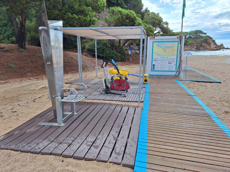 socorrista que acompanya a una nena amb una cadira que no és del tot amfíbia però que l'ajuda a estar a peu de sorra de la platja amb l'aigua just davant seu